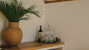 A room with a wooden shelf holding a potted plant, wine bottles, and glasses. There are towels on a lower shelf and a wicker basket on the floor. - Cabanas Blue (Mata de São João)