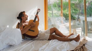 A person lying on a bed in a room with a bamboo ceiling, holding a guitar. - Cabanas Blue (Mata de São João)