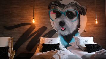 A hotel room with a large wall mural of a dog wearing glasses, a bed with white bedding, and a wooden headboard.