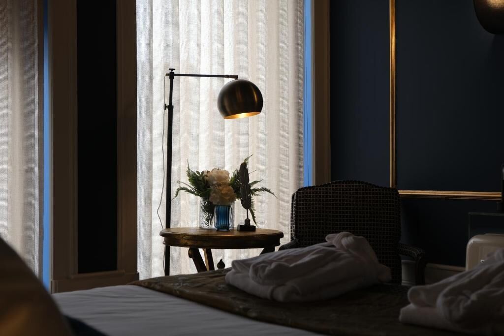 Una habitación de hotel con una cama, una mesita con un jarrón de flores, una silla y una lámpara de pie.