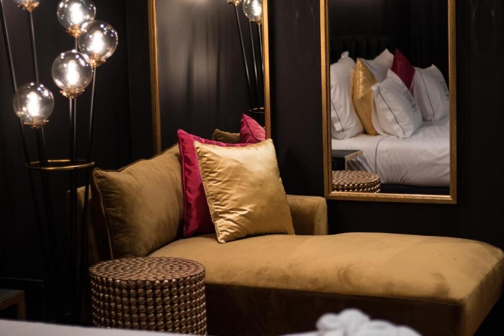 Una habitació acollidora amb un sofà, un otomà rodó i un mirall que reflecteix un llit amb almoades.