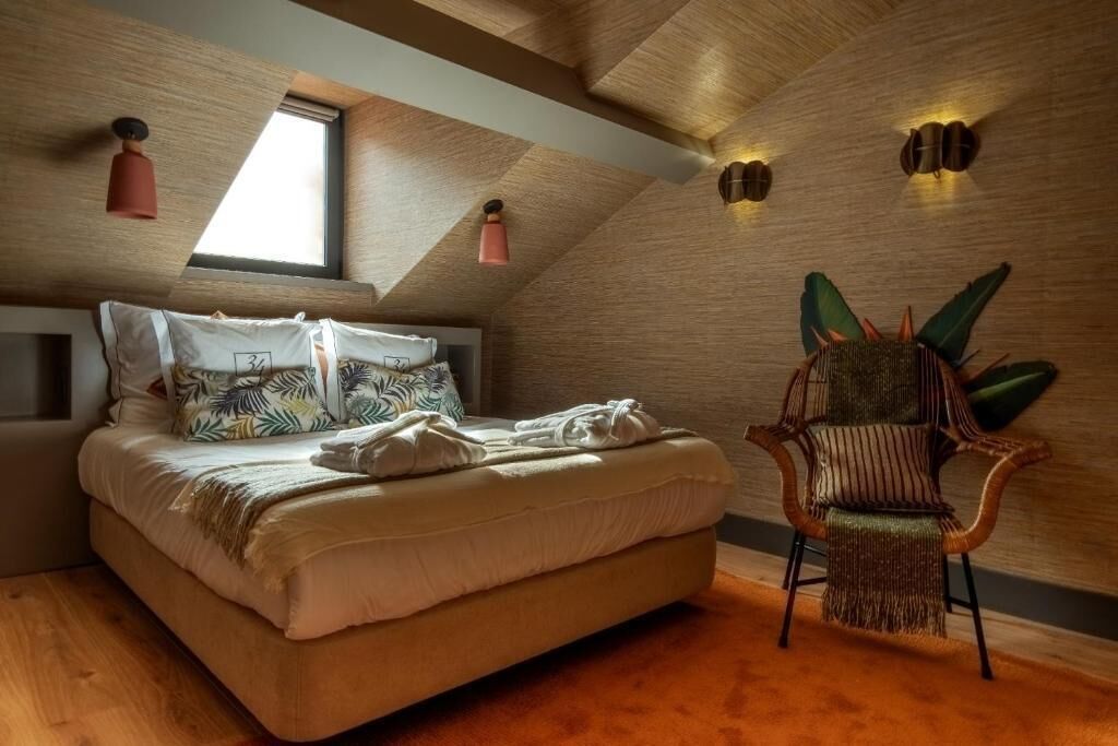 Slaapkamer met een schuine plafond, een bed met gekleurde kussens, een stoel met een groene kussen en wandlampen.