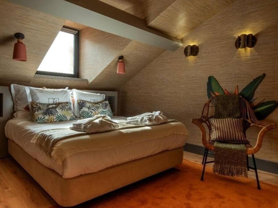 Slaapkamer met een schuine plafond, een bed met gekleurde kussens, een stoel met een groene kussen en wandlampen.