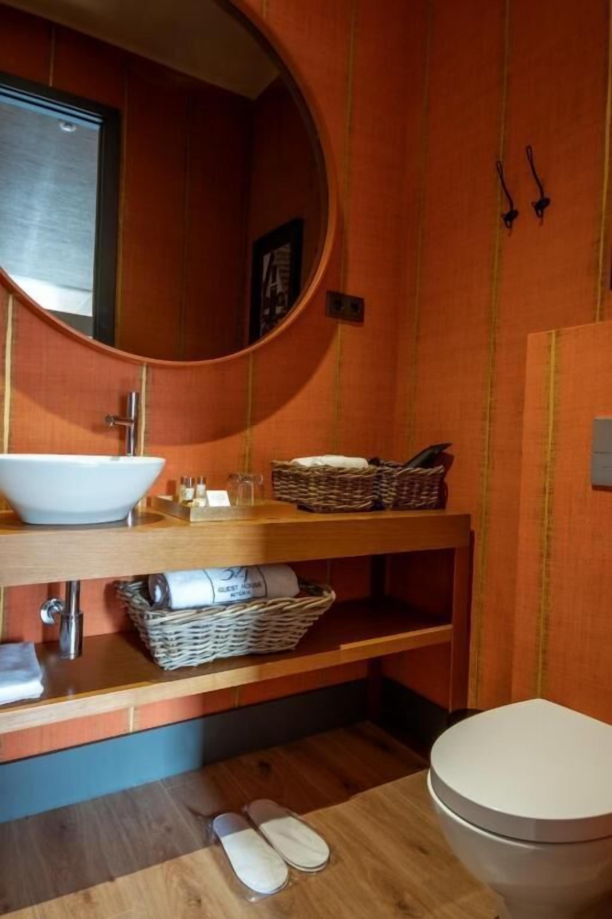 A bathroom with a round mirror, a white sink, a wooden vanity, a wicker basket, and a toilet.
