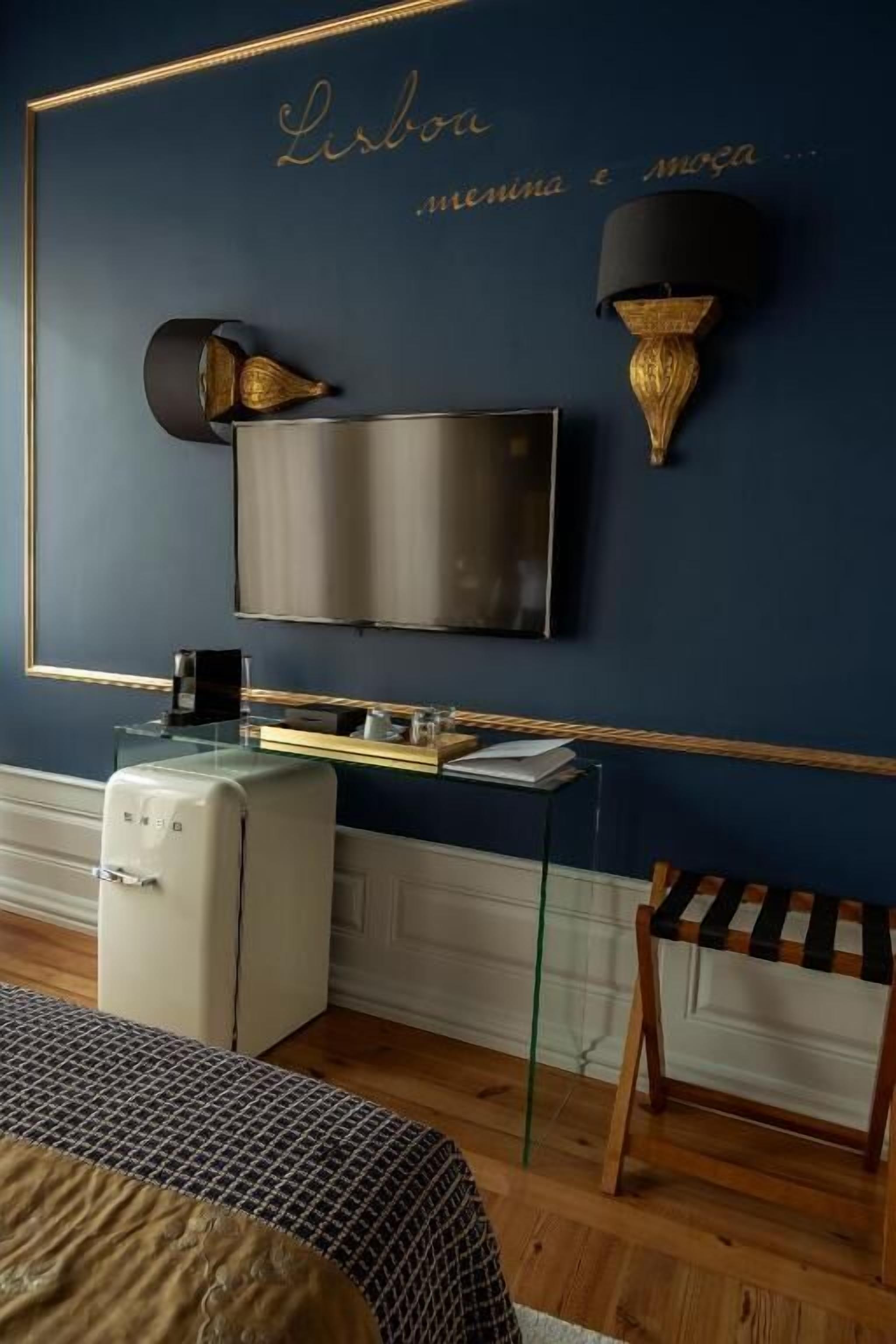 A room with a TV mounted on the wall, a small table with a lamp, and a bed with a patterned bedspread.