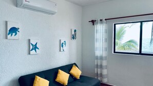 A modern living room with a dark green sofa, three yellow cushions, and a glass coffee table. There are four wall-mounted artworks depicting sea creatures. An air conditioning unit is mounted on the wall above the sofa. A window with a view of palm trees and the sea is visible, with a striped curtain partially drawn. - Beautifull Apartment beach front & nice sea view (Cancun)