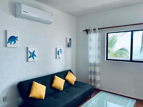 A modern living room with a dark green sofa, three yellow cushions, and a glass coffee table. There are four wall-mounted artworks depicting sea creatures. An air conditioning unit is mounted on the wall above the sofa. A window with a view of palm trees and the sea is visible, with a striped curtain partially drawn.