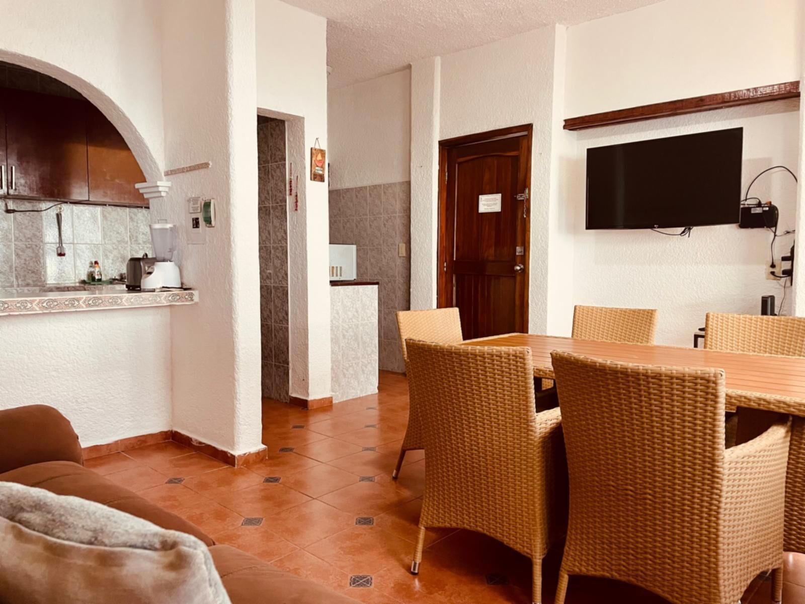 A dining area with wicker chairs and a wooden table, a kitchen with a tiled backsplash, and a flat-screen TV mounted on the wall.