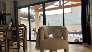 A cozy room with a large window, a dining table with chairs, and a plush armchair. - Maison "les Chênes D'hélios" en Drôme Provençale - Logement Entier (Réauville)