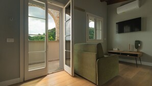An open sliding glass door leading to a balcony with a stone archway, a green upholstered chair, a flat-screen TV mounted on the wall, and a wooden floor. - Luce Boutique Hotel (Fortunago)