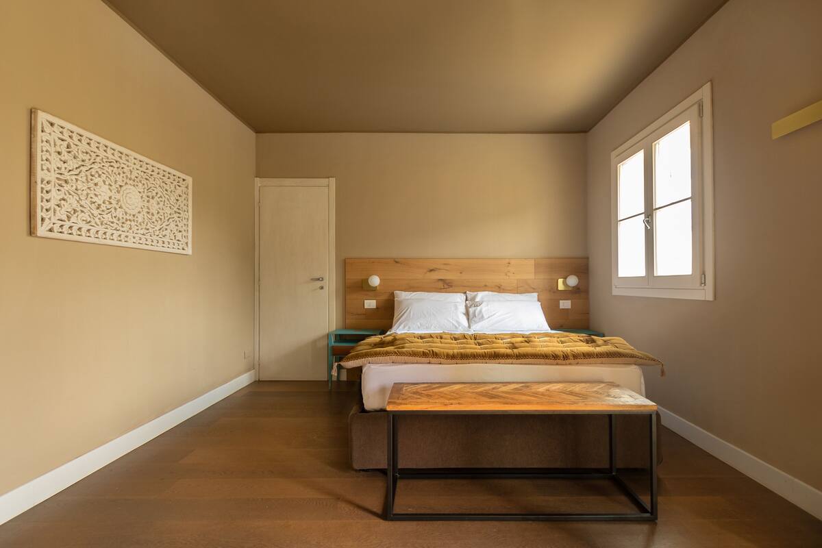 A bedroom with a bed, bedside tables, a wooden headboard, a decorative wall piece, and a window.
