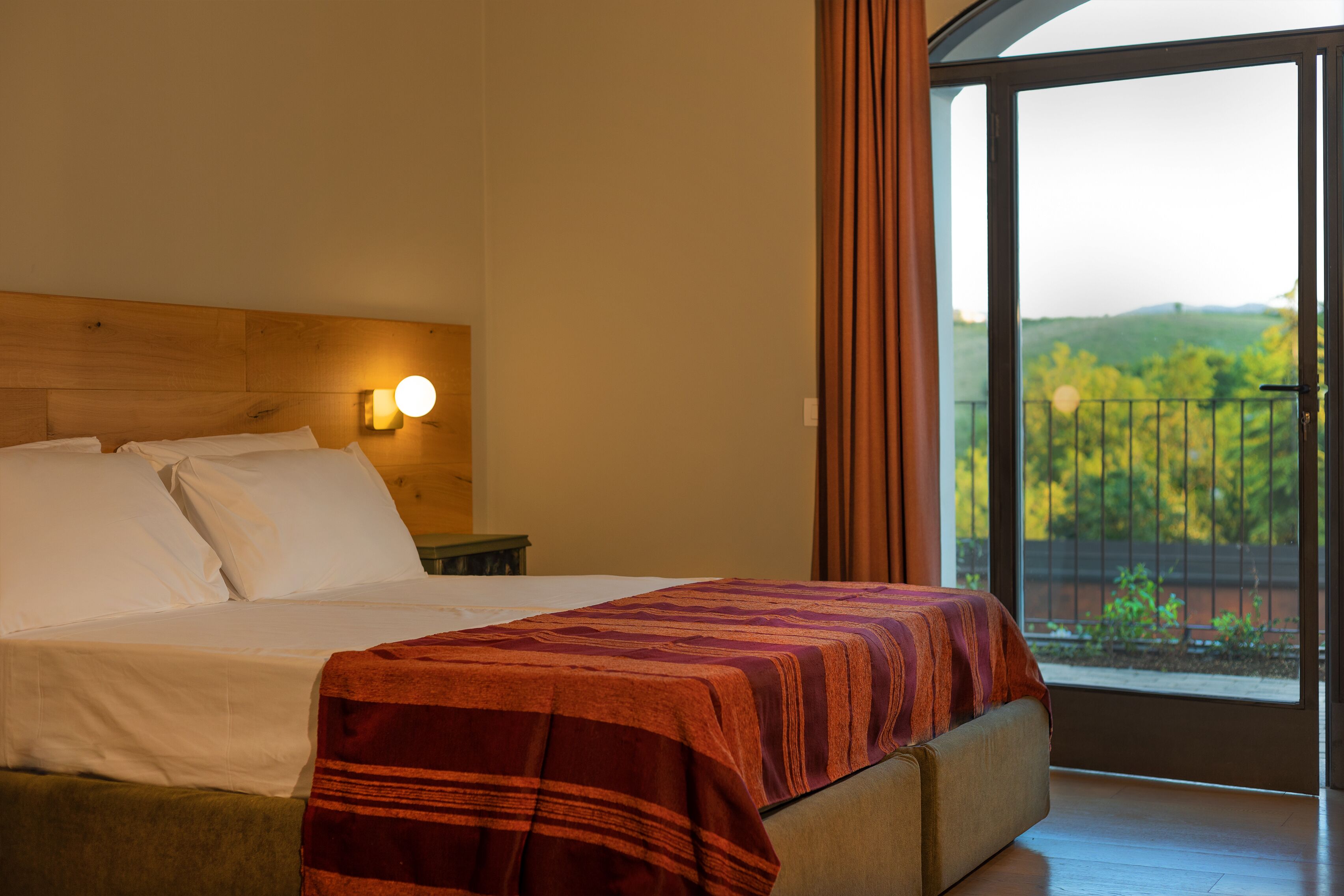 A hotel room with a bed, a nightstand, a wall-mounted lamp, and a large window with a view of greenery.