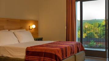 A hotel room with a bed, a nightstand, a wall-mounted lamp, and a large window with a view of greenery.
