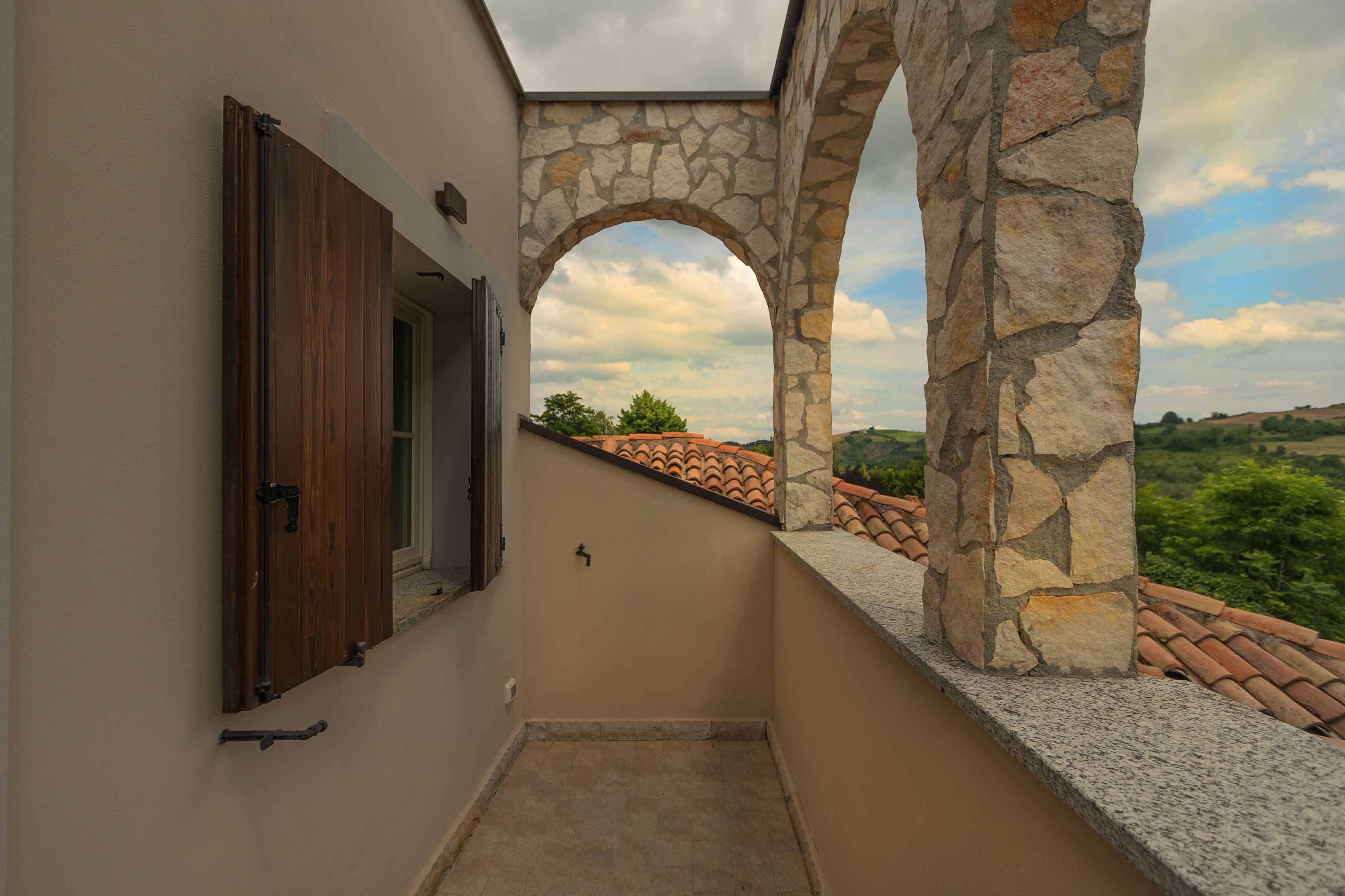 Un balcón estrecho con arcos de piedra, una puerta de madera y vistas a un tejado de tejas y vegetación.