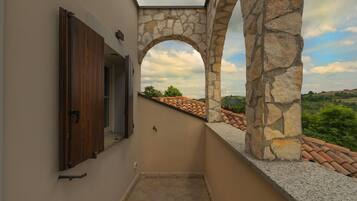 Un balcón estrecho con arcos de piedra, una puerta de madera y vistas a un tejado de tejas y vegetación.