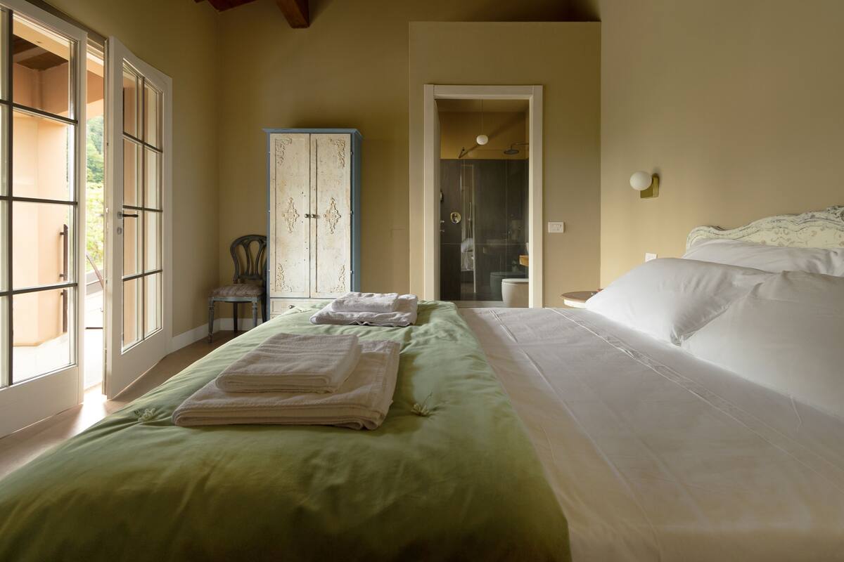 A bedroom with a bed, a chair, a wardrobe, and a bathroom visible through an open door.