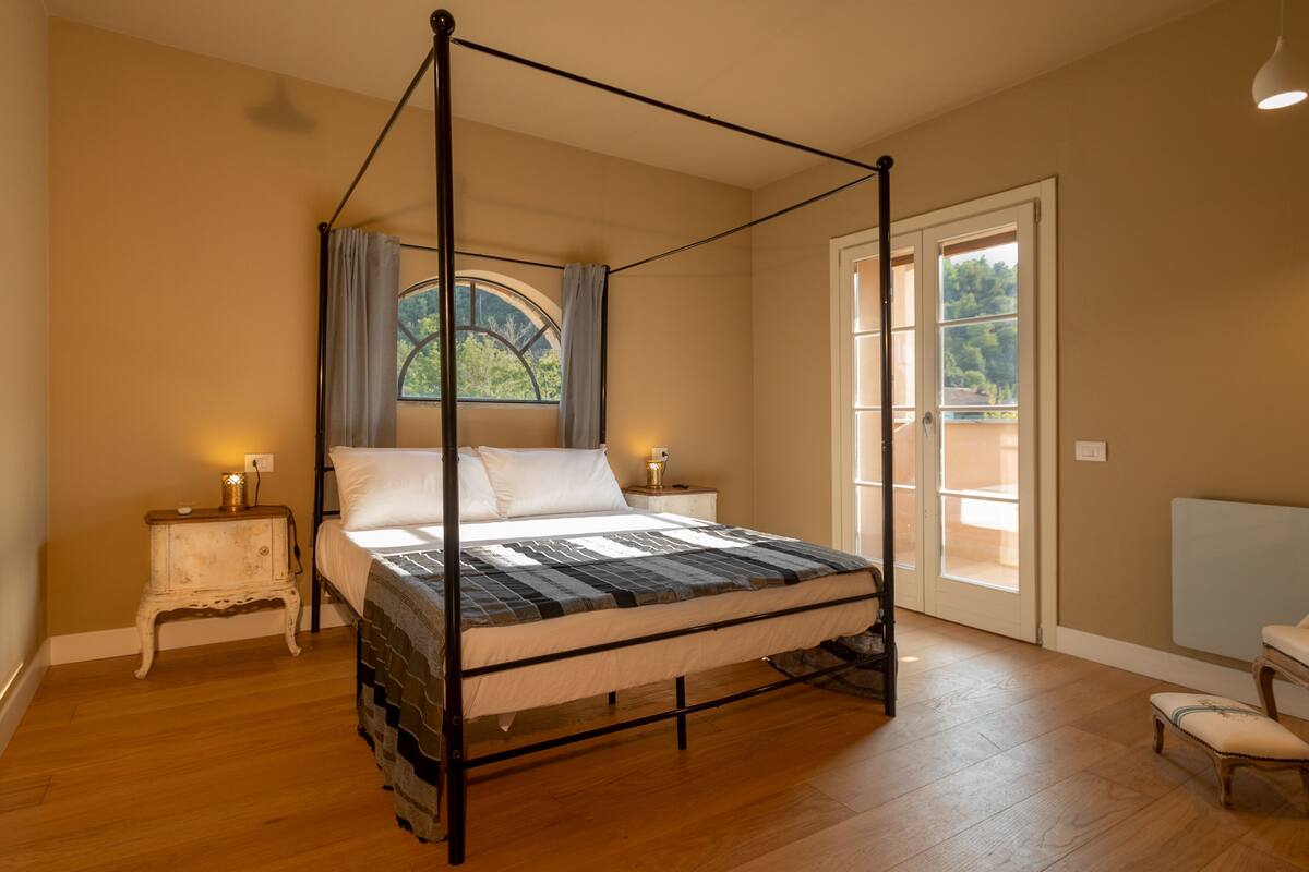 A four-poster bed with a view of the outdoors through a window, a small bedside table with a lamp, and a chair in the corner.