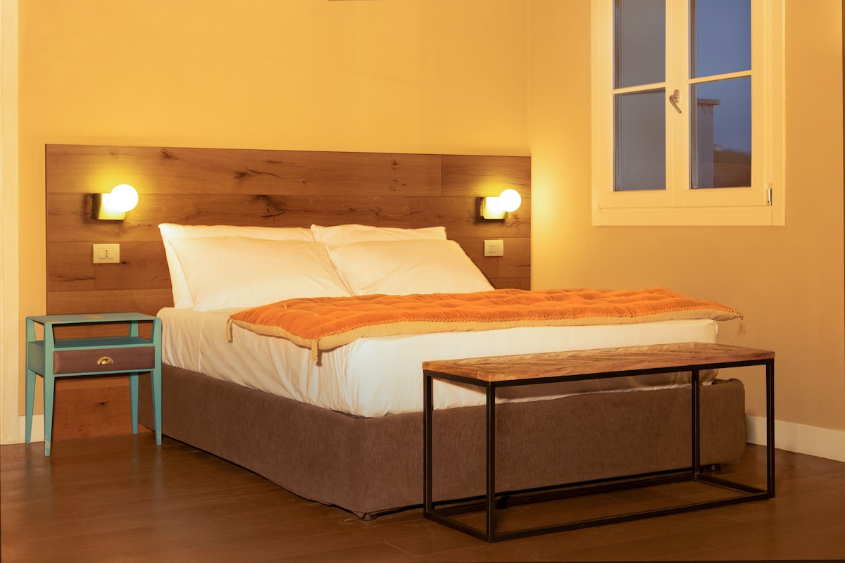 Una cama bien hecha con sábanas blancas y un plaid naranja, una mesita de noche de madera con un cajón azul verdoso, y una pequeña mesa con estructura metálica.