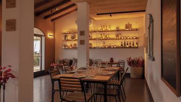 A dining area with a long table set for six, surrounded by chairs. There are shelves with bottles and glasses in the background.