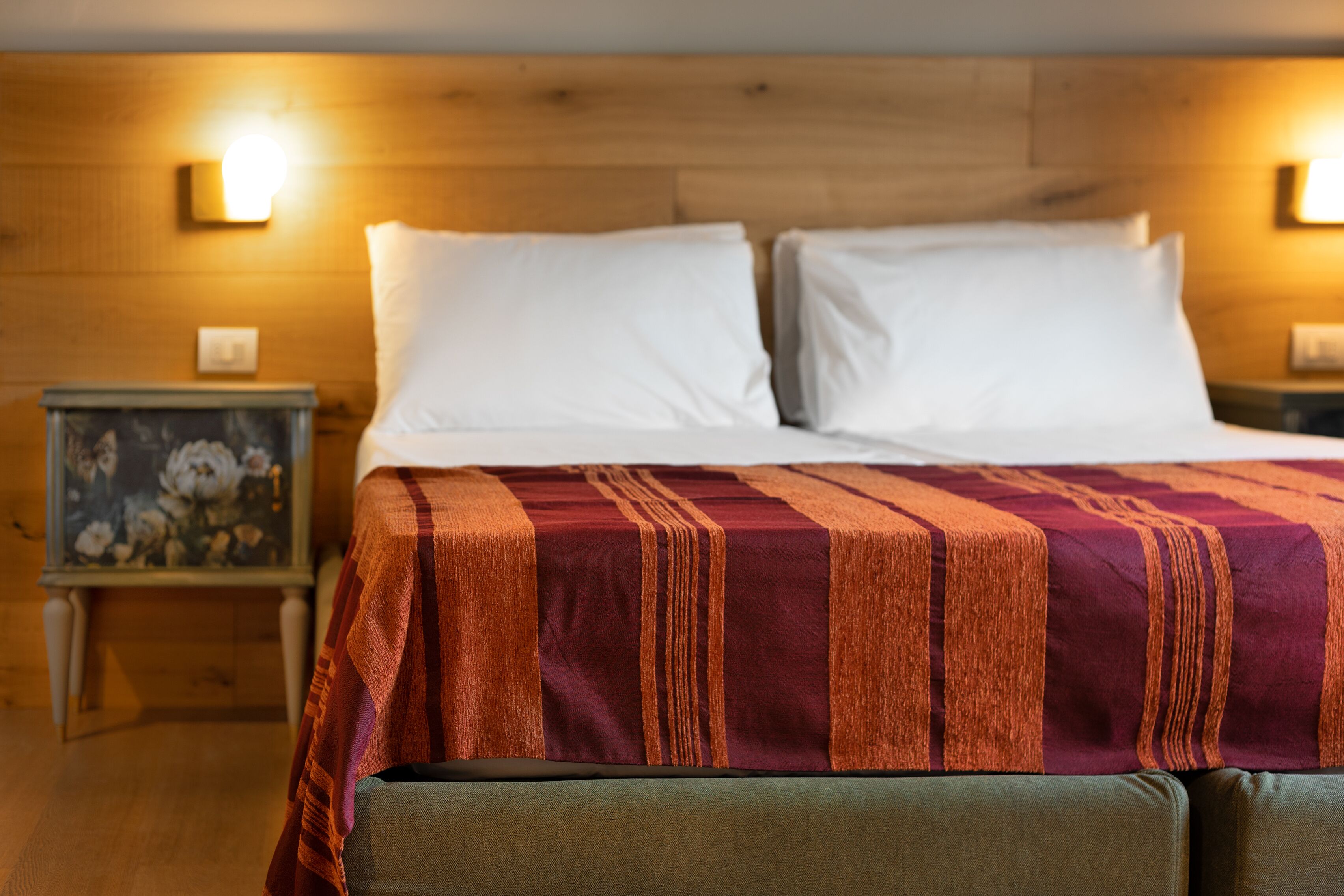 Una cama bien hecha con almohadas blancas y una colcha a rayas, un cabecero de madera y una mesita de noche con un cuadro enmarcado.