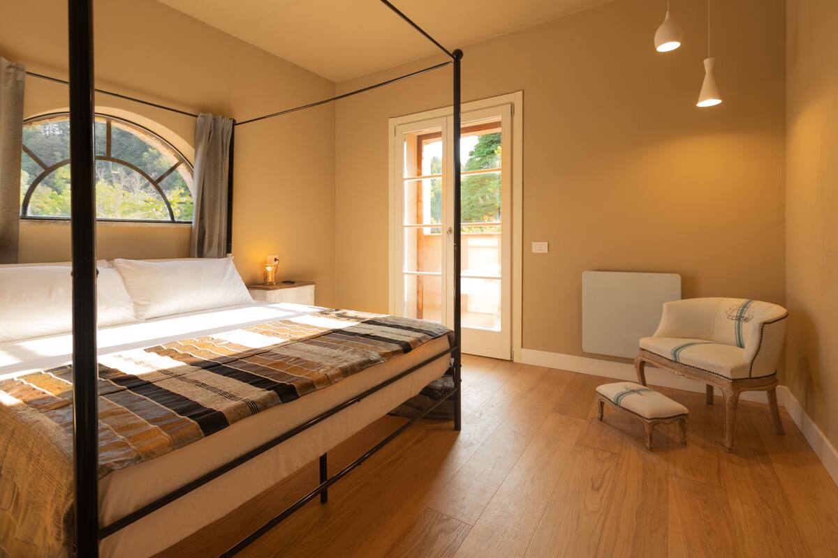 A bedroom with a four-poster bed, a chair, a small table, and a window with curtains.