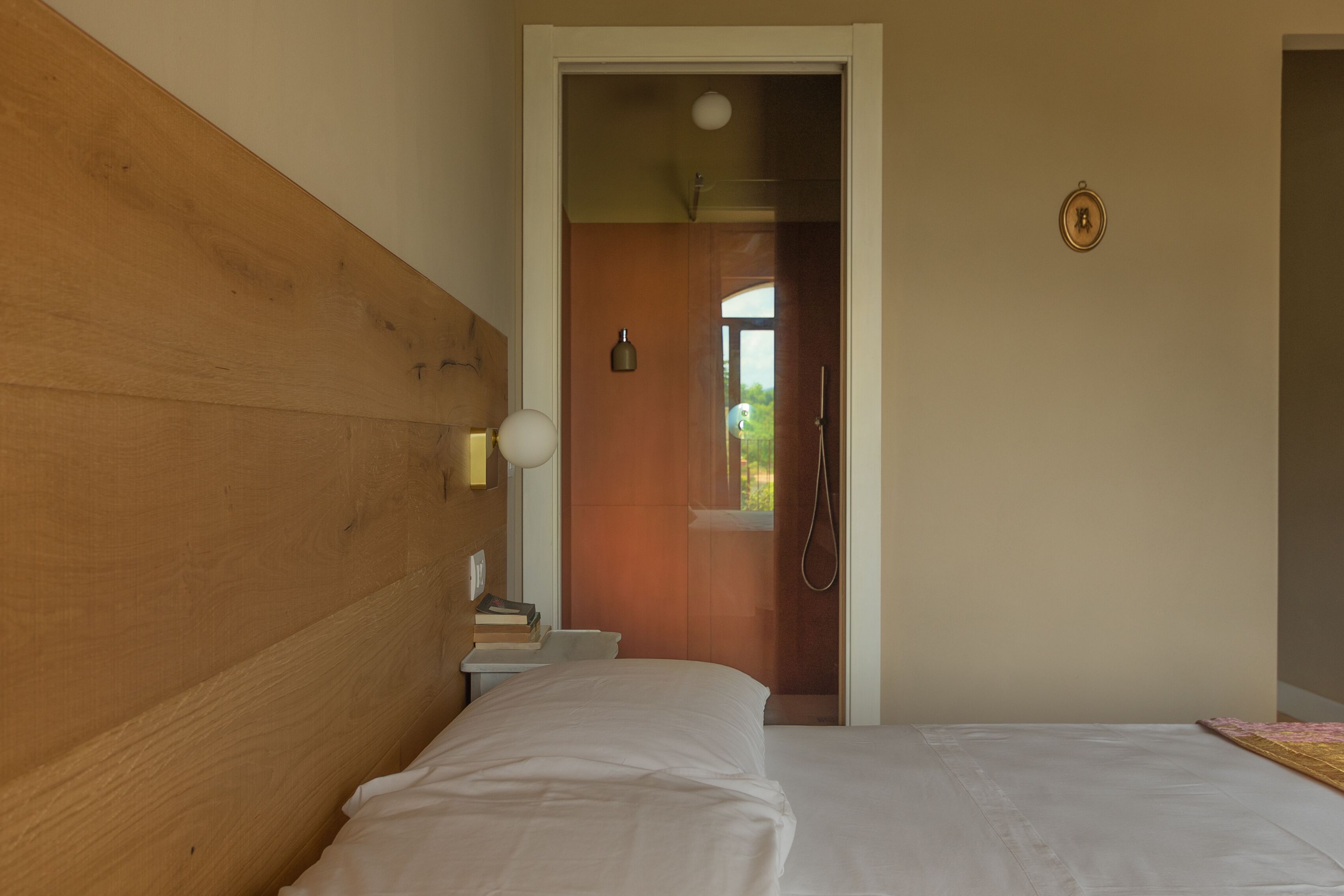 Um quarto com cabeceira de madeira, uma cama com roupa de cama branca, uma mesinha de cabeceira com abajur e uma porta que dá para um banheiro com chuveiro.