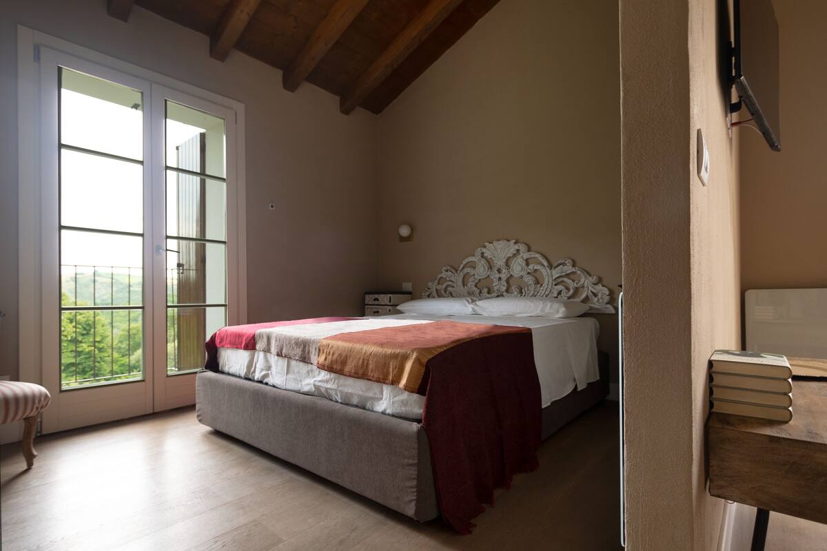 A bedroom with a large window, a bed with a headboard, a nightstand, and a wooden bench.