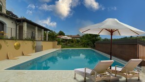 A swimming pool with lounge chairs and an umbrella, surrounded by a stone patio and a building with a tiled roof. - Luce Boutique Hotel (Fortunago)