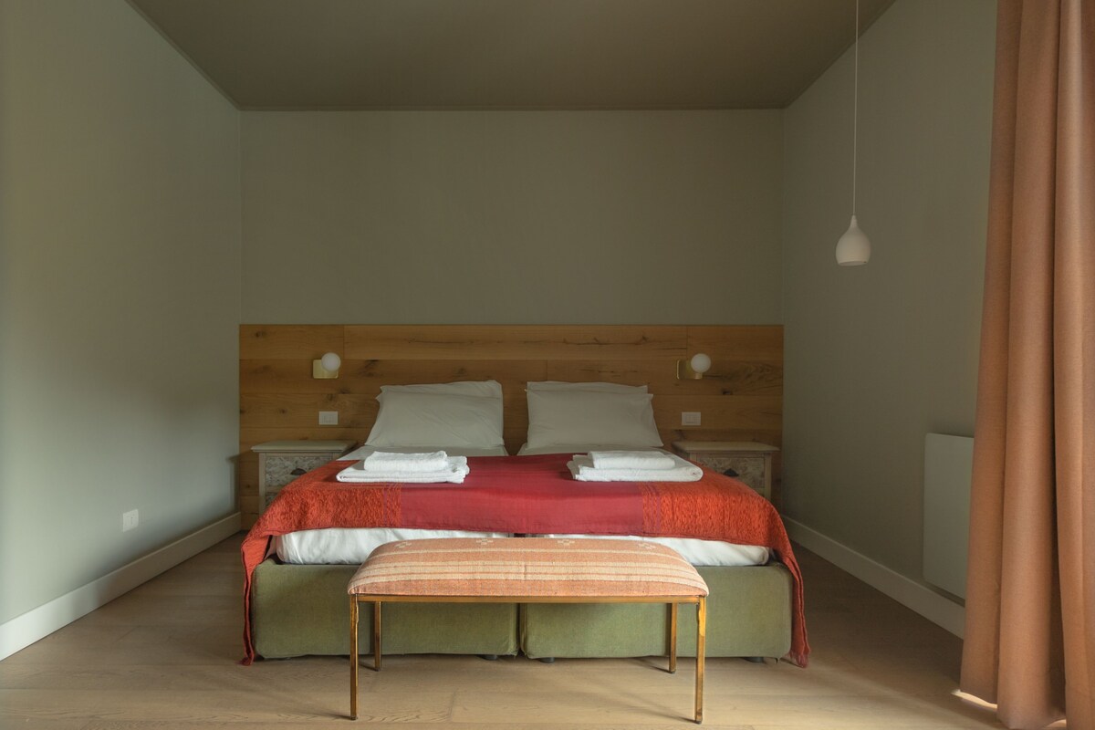 Un dormitorio con cabecera de madera, cama con ropa de cama roja y blanca, un banco y una mesita de noche con una lámpara.