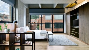 A modern bedroom with a large bed, a desk, and a TV. There is a view of buildings outside the window.