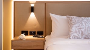 A hotel room with a neatly made bed, a bedside table with a phone and a remote control, and a wall-mounted light fixture.