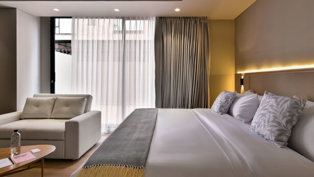 A modern hotel room with a bed, a sofa, a small table, and a window with curtains.