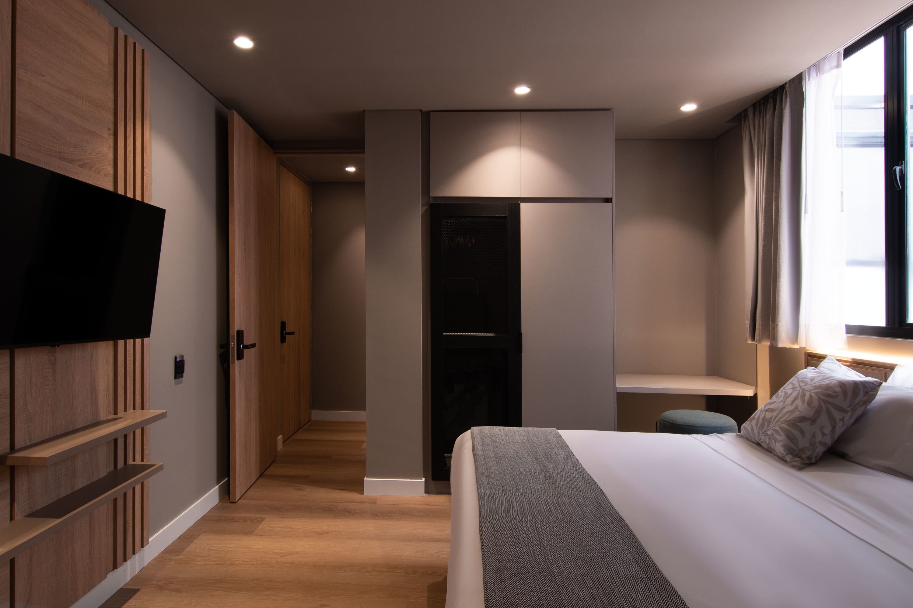 Une chambre d’hôtel moderne dotée d’un grand lit, d’un téléviseur à écran plat fixé sur un panneau en bois, d’un dressing et d’une fenêtre avec des rideaux.