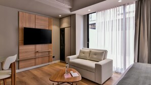 A modern hotel room with a flat-screen TV mounted on a wooden panel wall, a beige sofa, a wooden coffee table with a bottle and papers, and a large window with sheer curtains.
