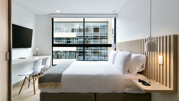 A modern hotel room with a large bed, a desk with a chair, a TV, and a view of the city through large windows. - w Magnificent Loft w City View in Santa Barbara (Bogotá)