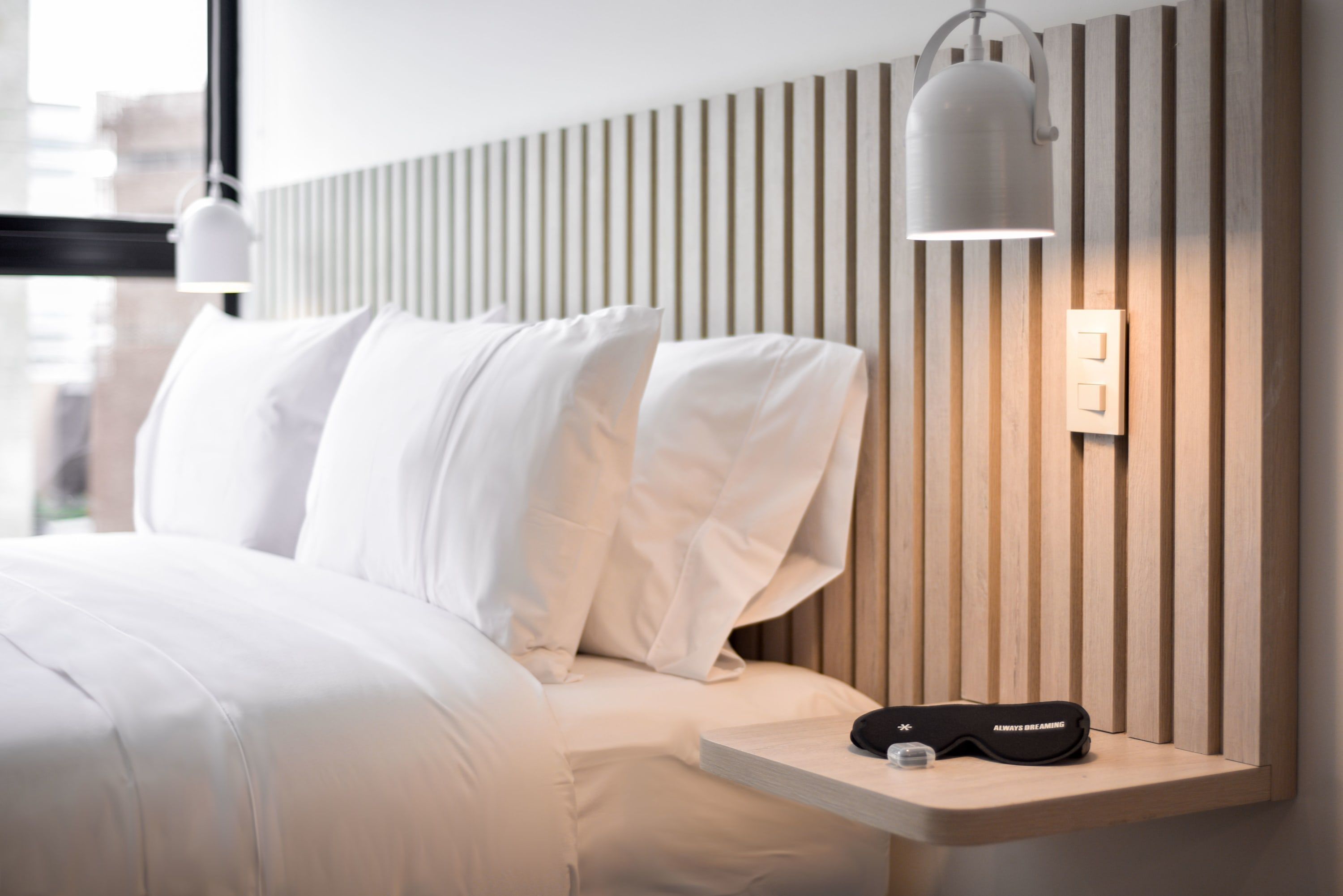 Una habitación de hotel moderna con una cama bien hecha, una mesita de noche con lámpara y una lámpara de pared.