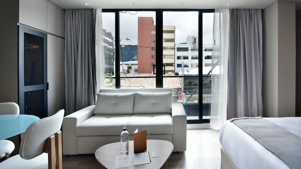 Habitació d'hotel moderna amb sofà, taula petita, llit i grans finestres que donen vistes a la ciutat.