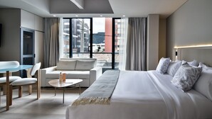 Habitació d'hotel moderna amb un llit gran, un sofà, una taula de menjador i una televisió. L'habitació té grans finestres amb vista a edificis i una muntanya.