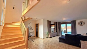 En rummelig opholdsstue med en trætrappe, en hængestol, en mørk sofa og et stort vindue med udsigt.