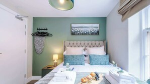 A neatly made bed with a patterned bedspread, a bedside table with a lamp, and a wall-mounted towel rack. - NEW! Stunning Studio in the City-Centre, Sleeps 2! (Chester)