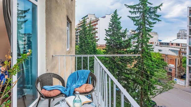 A balcony with wicker chairs, a table, and a glass railing overlooking a cityscape with trees and buildings. - HB apartments Mtskheta (Tbilisi)