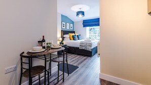 A compact hotel room with a bed, a small dining area with a table and chairs, and a window with blue curtains.