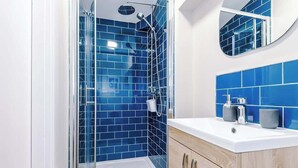 A modern bathroom with blue tiles, a glass shower enclosure, a white sink, and wooden cabinets.