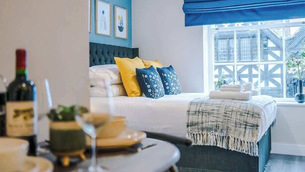 A hotel room with a bed, a window with blue curtains, a view of the outside, and a small table with a wine bottle and glasses.