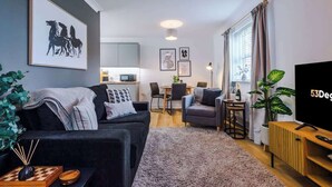 Un salon moderne avec un canapé, une table basse et un téléviseur. On y trouve également un coin repas avec une table et des chaises, et une cuisine à l’arrière-plan.