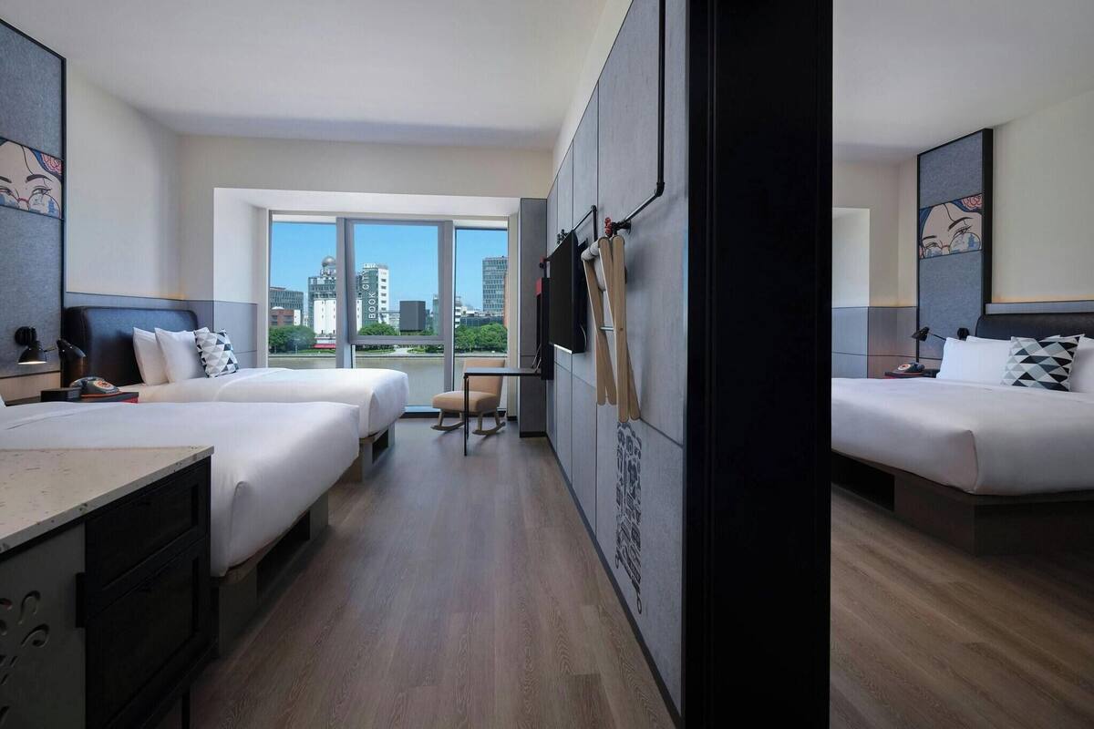A hotel room with two beds, a desk, a chair, and a large window offering a city view.