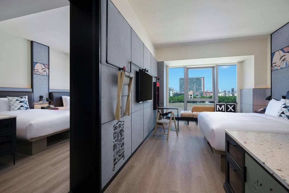 A hotel room with two beds, a desk, a chair, a TV, and large windows offering a city view.