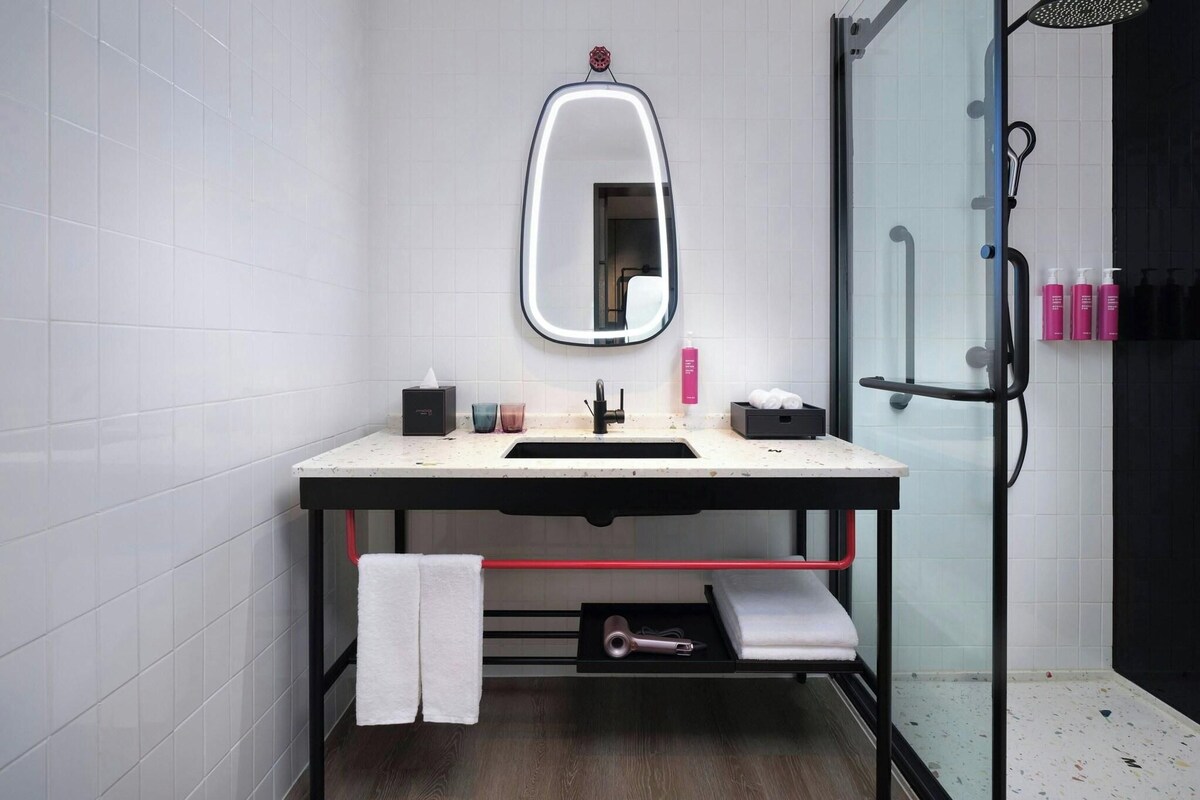 A modern bathroom with a white tiled wall, a large mirror, a sink with a countertop, a glass shower enclosure, and a towel rack with bottles.