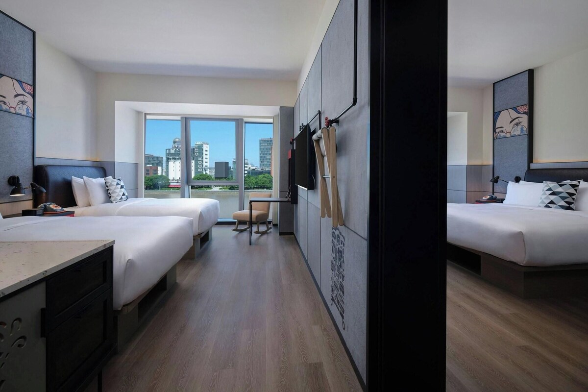 A hotel room with two beds, a desk, a chair, and a large window offering a city view.