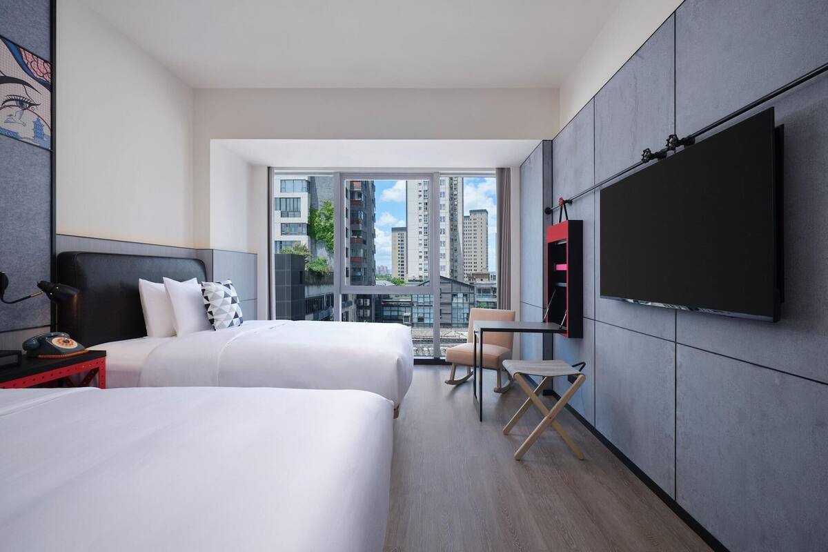 A modern hotel room with two beds, a TV mounted on the wall, a small table, and a chair.